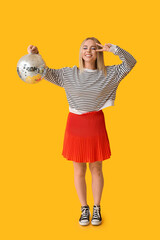 Poster - Beautiful young woman with disco ball showing victory gesture on yellow background