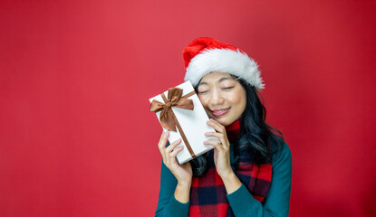 Wall Mural - Pretty smiling asian woman in warm christmas sweater and santa hat holding gift box as present with red background for season celebration