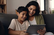 © AI_images - Smiling Latin mother with son using tablet at home, relaxing sitting on cozy couch together.