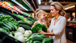 Stylish young woman mother standing near counters in green department in supermarket and smiling at child who sitting, mom holding fresh veg