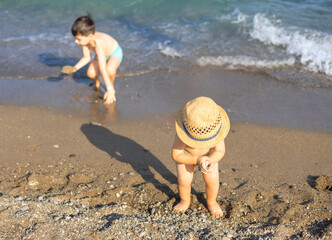 siblings 2 boys brothers wearing swimming trunks playing on beach summer vacation sunny day. kids wearing hat on head,digging in sands. sea waves in background warm weather.