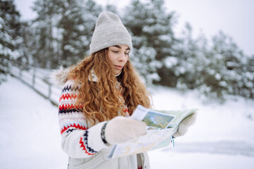 Curly-haired young woman standing in the wild with a map and compass in a snowy forest. Winter is approaching, the first snowfall. Adventure concept.