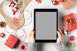 Tablet with a blank screen in a woman's hands surrounded by Christmas decorations and gifts, mockup for advertising