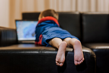 A small boy child in pajamas lies on a black leather sofa in the evening, watching a movie on a laptop online via the Internet. Legs, feet close-up. Happy childhood, lifestyle.