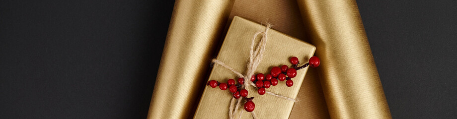 Canvas Print - shiny gift box with red holly berries on golden paper and black backdrop, horizontal banner