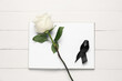 Rose with black ribbon and blank open notebook on white wooden background