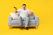 © Pixel-Shot - Surprised young man sitting on sofa and watching TV with popcorn against yellow background