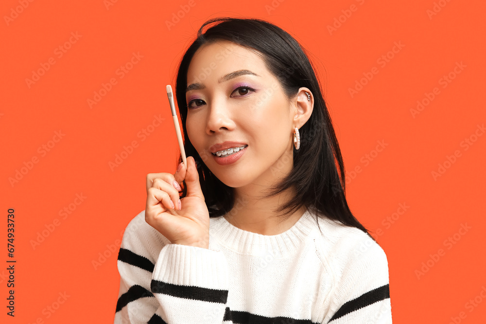 Young Asian woman with makeup brush on orange background