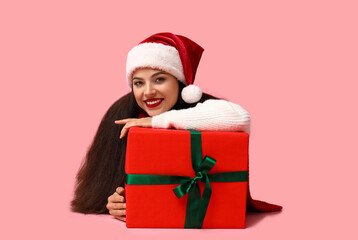 Beautiful young woman in Santa hat with Christmas gift on pink background