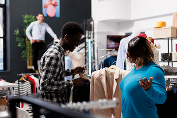 Canvas Print - Stylish client shopping for fashion collection, looking at shirt fabric in modern boutique. African american couple buying fashionable clothes, checking hangers with formal wear in clothing store