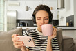 Portrait of woman in her living room, sitting on sofa and watching videos on mobile phone, wearing headphones, drinking tea or coffee in pink cup