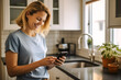 © MVProductions - A happy caucasian woman standing in her kitchen white looking at good news on her smartphone