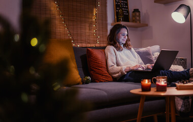 A young woman spends a cozy winter evening at home lying on the sofa with a laptop. Winter holidays, Christmas and online surfing concept