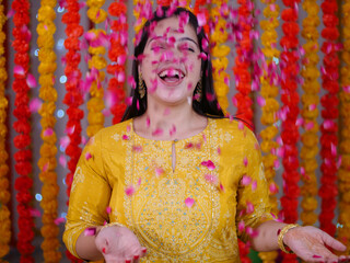 Shot of a beautiful Indian female smiling while throwing flower petals - Festive vibe  newly wed woman. Happy young girl laughing and looking towards the camera in traditional wear - festival backg...