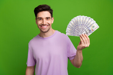 Poster - Photo of successful man with brunet hair dressed purple t-shirt presenting bunch of dollars in arm isolated on green color background