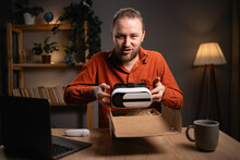Man unpacking parcel with VR Glasses, looking inside, sitting at home desk, customer opening cardboard box with online store order and recording unboxing video, good shipping delivery