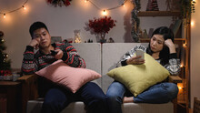 bored Japanese man propping face and surfing channels with a tv control while his wife is using phone, ignoring each other on the sofa on Christmas night in the living room at home