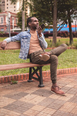 Young black man, sitting on the bench in the square, talking on his smartphone.