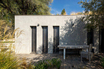 Wall Mural - Front view of a contemporary concrete house with large windows, a table, and a chair surrounded by green environment.