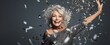 Happy beautiful senior mature woman in luxury dress celebtraing New Years Eve party or birthday, event. Stylish grey hair senior exited woman with glitter confetti celebrating new year