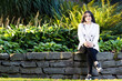 Young beautiful lady poses sitting at a park with beautiful garden. She is wearing casual with white blazer and blouse and black jeans