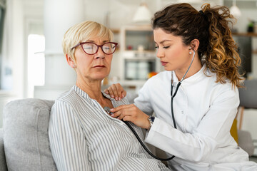 Wall Mural - Serious female doctor examining senior woman with stethoscope while sitting on sofa at nursing home.