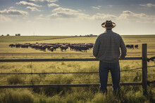 The Back View Of A Big Man In Cowboy Wide Brimmed Hat And Boots Stand Near A Wooden Fence While Looking At The Cow Herd In The Field. Generative AI.