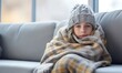 © Eli Berr - Toddler girl wear hat wrapped in plaid sit alone shivering from cold on sofa, heated apartment without heating due debt. Unhealthy child feeling discomfort try to warming up