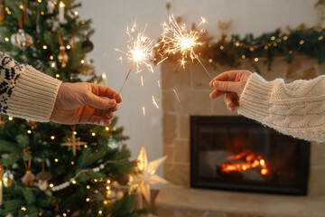 Wall Mural - Happy New Year! Hands holding burning fireworks against modern fireplace and christmas tree with golden lights. Friends and family celebrating with burning sparklers in hands, atmospheric eve