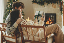 Happy Couple In Cozy Sweaters Exchanging Stylish Wrapped Christmas Gifts On Background Of Fireplace With Festive Mantle And Modern Decorated Christmas Tree With Lights. Merry Christmas!