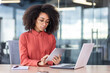 © Liubomir - Serious confident woman working with tablet computer inside office, business woman thinking sitting at desk with laptop, business woman in shirt and curly hair