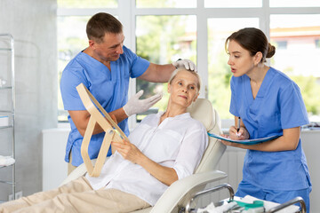 Elderly woman holding mirror while skilled male cosmetologist showing results of facial procedure in medical aesthetic office. Female assistant filling out patient medical record