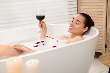 Canvas Print - Happy woman with glass of wine taking bath in tub with foam and rose petals indoors