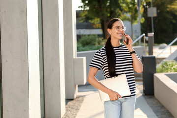 Canvas Print - Happy young woman with modern laptop and talking on smartphone outdoors. Space for text