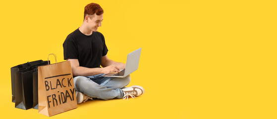 Happy young redhead man with shopping bags and laptop sitting on yellow background with space for text. Black Friday sale