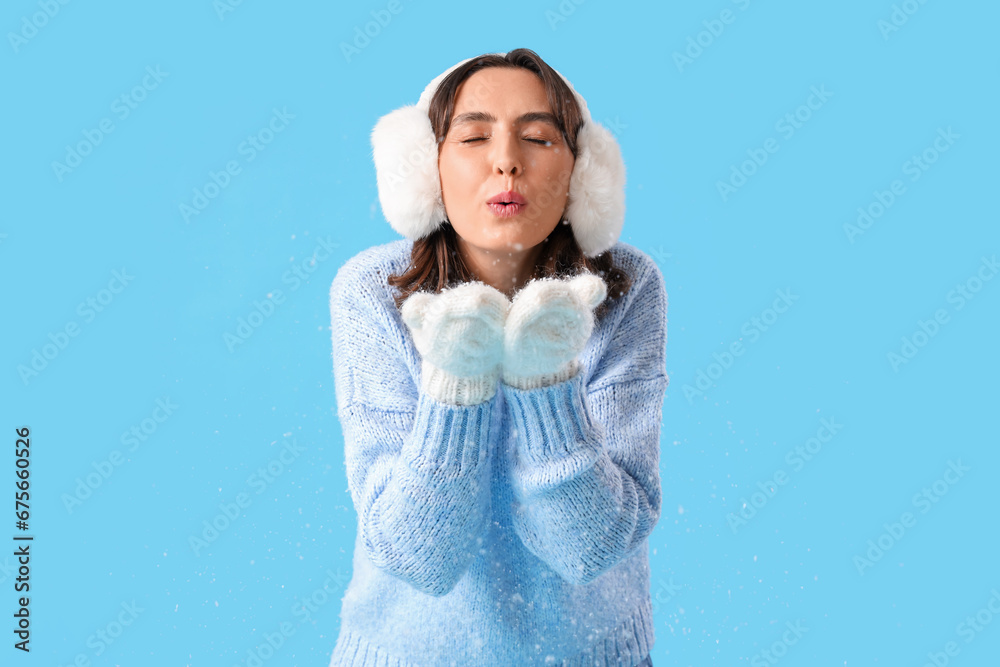 Young woman in warm clothes blowing snow on blue background
