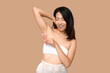 Young Asian woman with razor shaving underarms on brown background