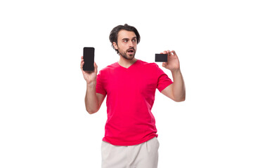 Wall Mural - young surprised male promoter dressed in a red basic t-shirt holds a plastic card and a mobile phone