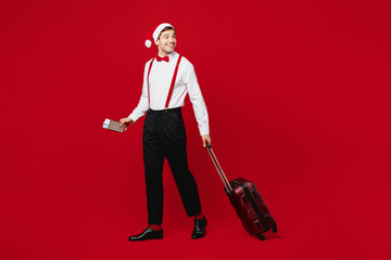 Poster - Full body traveler man wear Santa hat posing hold suitcase passport ticket look aside isolated on plain red background. Tourist travel abroad in free time rest getaway Air flight trip journey concept