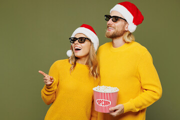 Wall Mural - Merry young couple two friends man woman wear 3d glasses sweater Santa hat posing watch movie film eat popcorn in cinema point aside isolated on plain green background. Happy New Year holiday concept.