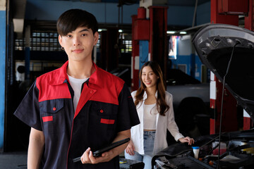 Portrait of young Asian male automotive mechanic worker with female customer's EV car at maintenance garage, happy smiles, professional service station, check and repair in auto transport industry.