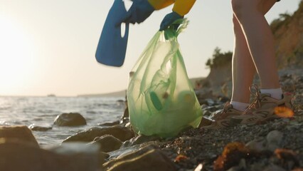 Wall Mural - Low angle view of woman volunteer picking up bottle and put it in garbage bag on wild pebble coast. Concept of environmental pollution