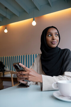 Smiling young woman in hijab using phone in cafe