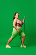 Motivated young overweigh woman in sportswear training, doing exercises with fitness expanders against green studio background. Concept of sport, body-positivity, weight loss, body and health care