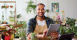 © serg - Portrait of african american man using digital tablet computer sitting wooden desk with botanic plants and looking into camera smiling. Male seller working at floral store checking order list on