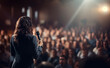 Rear view of motivational woman speaker standing on stage in front of audience for motivation speech on business event