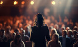 Fototapeta  - Rear view of motivational woman speaker standing on stage in front of audience for motivation speech on business event