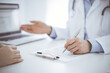 © rogerphoto - Doctor and patient sitting opposite each other at the desk in clinic. The focus is on female physician's hands filling up the medication history record form or checklist, close up. Medicine concept