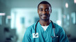 Dark-skinned afro american young male doctor in medical clothing smiles happily and looks at camera. Blurred background of the hospital, office in background. Healthcare concept, medicine