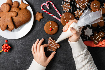 Wall Mural - baking, cooking, christmas and food concept - close up of hands with pastry bag decorating gingerbread cookies with white icing on black table top